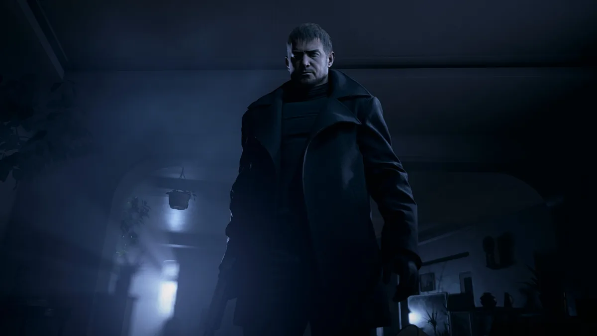 Chris Redfield en long manteau sombre debout dans une pièce éclairée par une lampe dans Resident Evil Village